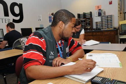 A tutor sitting at a desk, working from a textbook with a notebook and calculator.