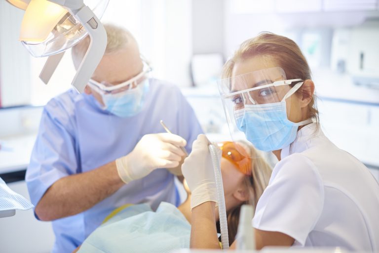 A dental assistant turns and smiles to camera during a dental procedure. She is wearing a mask, protective eye shield and is holding the suction hose. Behind her the senior dentist can be seen checking the patients dodgy tooth. The brightly lit dentist surgery is clean and modern.