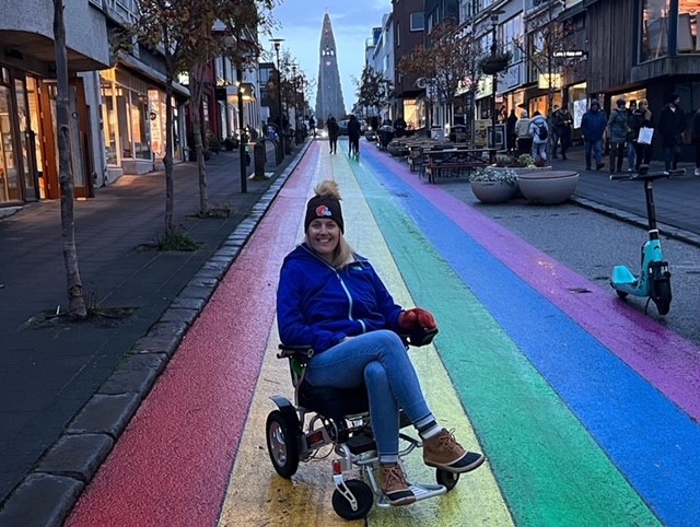 Elizabeth "Biz" Them sits in her wheelchair on a rainbow painted street